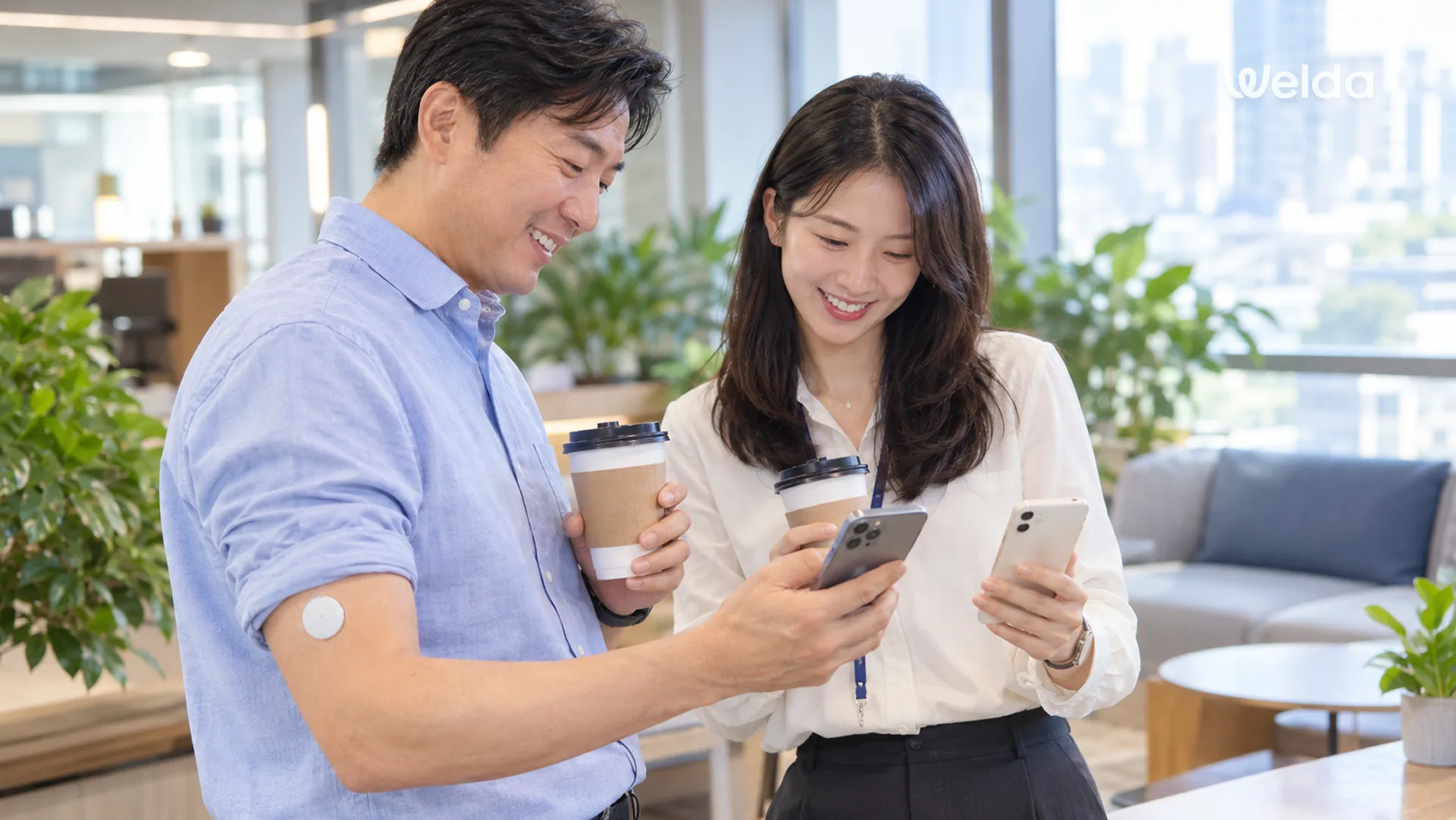 사무실 라운지에서 팔에 연속혈당측정기 센서를 붙인 임직원 두 명이 커피를 들고 스마트폰 앱으로 혈당 변화를 함께 확인하는 모습
